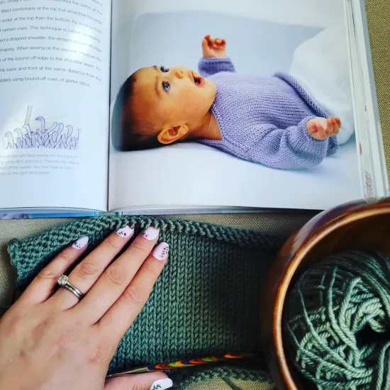 a green baby sweater knitting project in progress. a page of from Debbie Bliss's book Baby Knit for Beginners is also displayed.