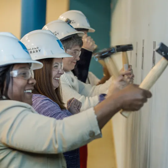 Four staff members wearing white hard hats swing hammers into a wall during a wall breaking ceremony