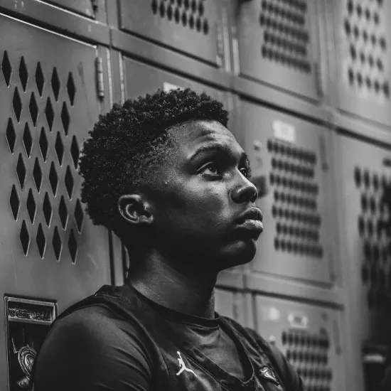 Black and white image of a Black young man.  He is leaning against lockers and wears a basketball jersey.  His hair is short and he stares focused on the game ahead.