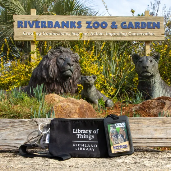 A Riverbanks Zoo and Garden entrance sign stands behind bronze lion statues surrounded by greenery. In the foreground, a Richland Library “Library of Things” pouch and a Riverbanks Zoo and Garden community pass rest on a wooden ledge.