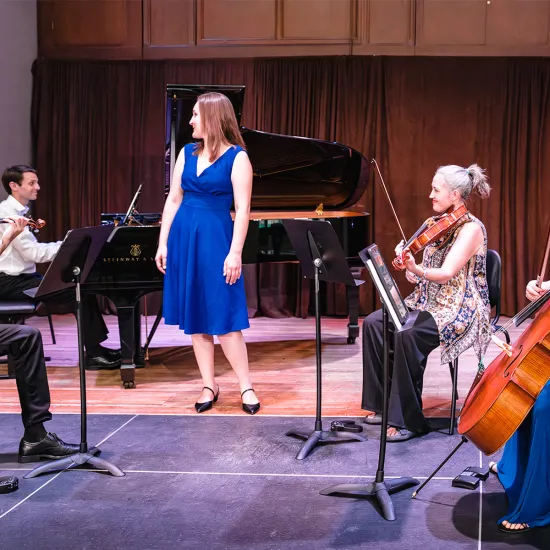 Members of Some Strings Attached perform string instruments on stage. Four members are sitting while singer Brittany Martin stands in the center wearing a blue dress.